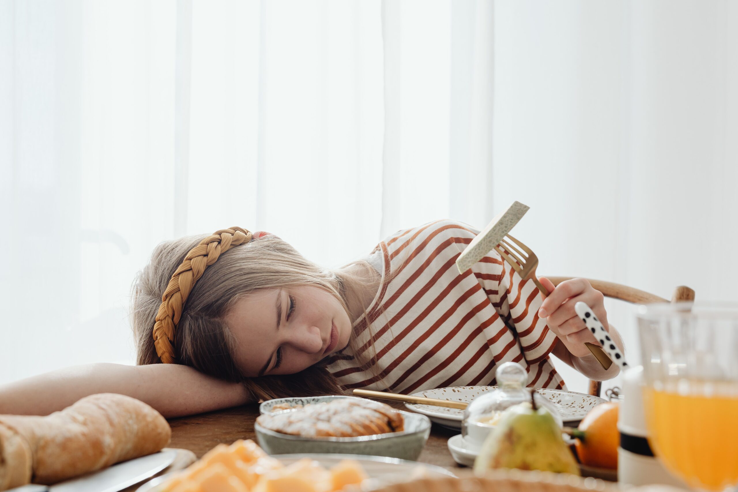 Trött flicka med flätat hårband vilar huvudet på bordet vid frukosten